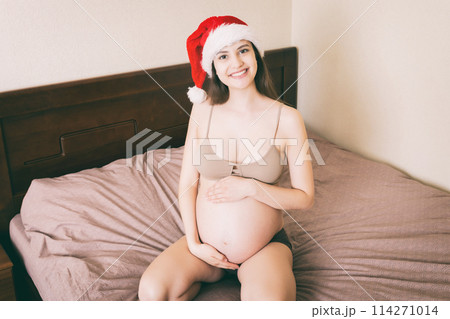 Cute happy pregnant woman in pajamas at home in santa hat. Mother to be with big belly, hands over tummy. Merry Christmas holidays. New Year Eve concept 114271014