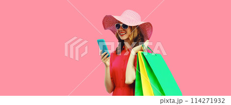 Beautiful happy young woman looking at mobile phone with colorful shopping bag in summer hat on pink 114271932