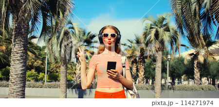 Happy smiling young woman listening to music in headphones with phone in summer park with palm trees 114271974