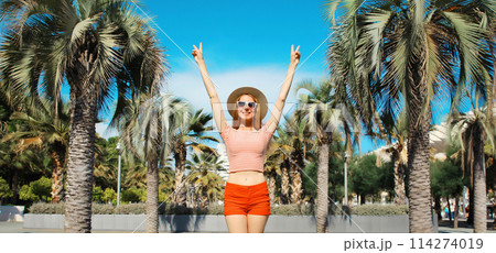 Summer vacation, happy cheerful young woman raising hands up against a palm trees background 114274019