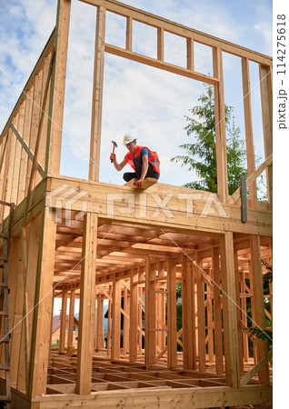 Wooden two-story frame house being built by carpenter. Man wearing glasses hammering nails using hammer, dressed in protective helmet and construction vest. Concept of modern ecological construction. 114275618