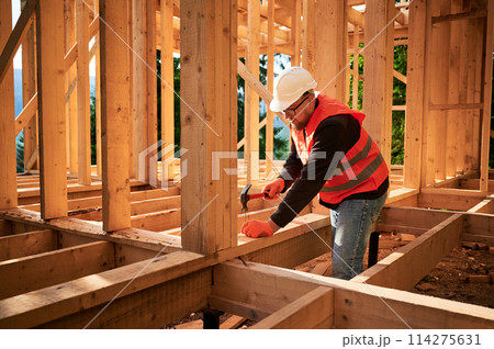 Carpenter constructing wooden frame house. Bearded man with glasses is hammering nails into the structure, wearing protective helmet and construction vest. Concept of modern ecological construction. 114275631