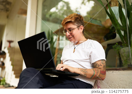 Tattooed Gen Z transgender person works on laptop in cozy office space, displays casual, inclusive workspace. Focused yet comfortable, represents modern work-life balance, LGBTQ-friendly environment. Tattooed Gen Z transgender person works on laptop in cozy office space, displays casual, inclusive workspace. Focused yet comfortable, represents modern work-life balance, LGBTQ-friendly environment. 114275775