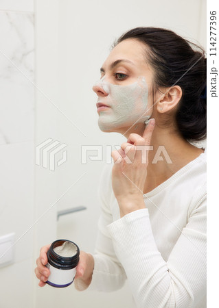 Caucasian woman looking in mirror and applying face mask white in bathroom Caucasian woman looking in mirror and applying face mask white in bathroom 114277396