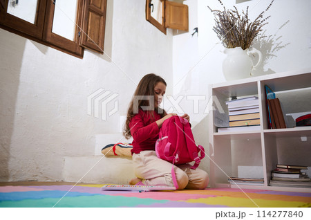 Caucasian beautiful little kid girl, elementary age school child packing her backpack, sitting on colorful puzzle carpet at cozy home interior. The concept of Back to school 114277840