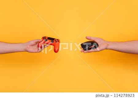 In one hand, a gamepad, and in the other hand, a computer mouse, reaching towards each other against a yellow background In one hand, a gamepad, and in the other hand, a computer mouse, reaching towards each other against a yellow background 114278923