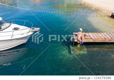 Cute little girl kid sits on wooden pier next moored boat yacht with scenic turquoise sea water background. Happy child in cap hold fishing net have fun explore nature at summer vacation 114279166