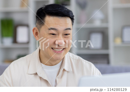 A middle-aged Asian man showing a joyful expression while interacting on his laptop in a stylishly decorated living room. Perfect imagery for themes on remote work, happiness, and technology use at A middle-aged Asian man showing a joyful expression while interacting on his laptop in a stylishly decorated living room. Perfect imagery for themes on remote work, happiness, and technology use at 114280129