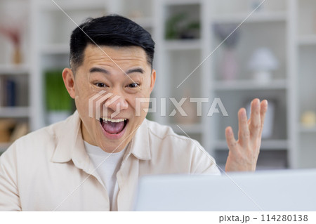 An enthusiastic Asian adult man expressing joy and success while working from home. He is visibly cheerful, gesturing with his hands, in a brightly lit room. An enthusiastic Asian adult man expressing joy and success while working from home. He is visibly cheerful, gesturing with his hands, in a brightly lit room. 114280138