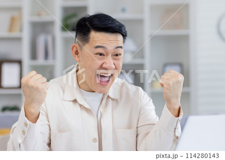 Middle-aged Asian man in a cream shirt joyously celebrating while looking at his laptop screen, evidently thrilled by good news or success at home. Middle-aged Asian man in a cream shirt joyously celebrating while looking at his laptop screen, evidently thrilled by good news or success at home. 114280143