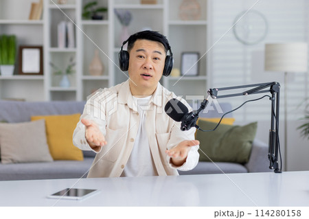 An engaging Asian man talking and looking at the camera while recording a podcast in a home studio setting. 114280158