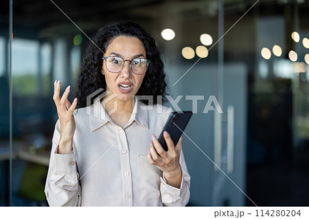 A businesswoman appears upset and frustrated while holding a smartphone in a contemporary office setting. Her expression conveys a sense of urgency and dissatisfaction. 114280204
