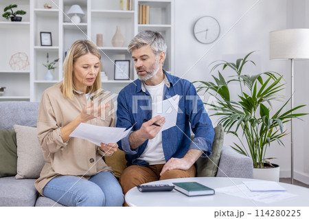 A mature couple sits closely on a sofa in a well-appointed living room, discussing documents with concern and concentration. A mature couple sits closely on a sofa in a well-appointed living room, discussing documents with concern and concentration. 114280225
