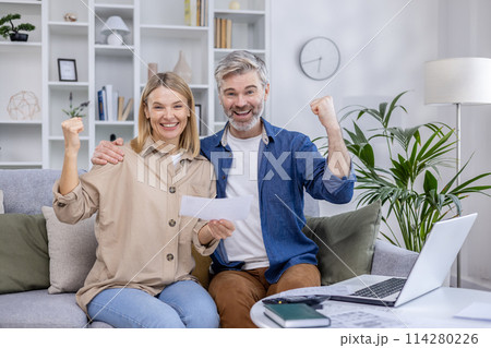 Middle-aged couple celebrating while looking at their laptop in a modern living room, showing joy and excitement over positive news. Middle-aged couple celebrating while looking at their laptop in a modern living room, showing joy and excitement over positive news. 114280226