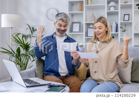 A mature couple expresses joy while looking at a document in their living room, conveying excitement and happiness. A mature couple expresses joy while looking at a document in their living room, conveying excitement and happiness. 114280229