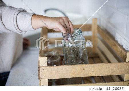 Sorting glass for recycling. A woman in the kitchen carefully places a glass jar into a recycling bin. A woman puts glassware into a recycling bin. Glass sorting concept. 114280274