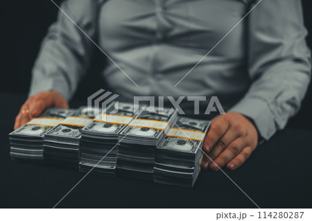 Neatly folded stacks of hundred dollar bills on a black background in front of a man. Close-up. On his sides are beautiful stacks of dollars in bundles. Big money concept. 114280287