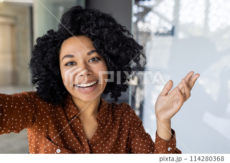 A cheerful young woman in a polka dot shirt takes a selfie, expressing joy and confidence in a modern indoor setting with natural light. A cheerful young woman in a polka dot shirt takes a selfie, expressing joy and confidence in a modern indoor setting with natural light. 114280368