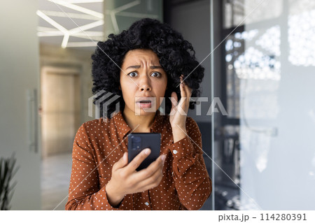A young woman in a polka dot shirt looks shocked and upset while reading bad news on her smartphone in a modern office setting. A young woman in a polka dot shirt looks shocked and upset while reading bad news on her smartphone in a modern office setting. 114280391
