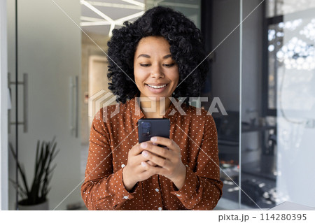 A cheerful young woman stands in a stylish office, engrossed in her smartphone. Her engagement conveys connectivity and professionalism in a relaxed corporate environment. A cheerful young woman stands in a stylish office, engrossed in her smartphone. Her engagement conveys connectivity and professionalism in a relaxed corporate environment. 114280395