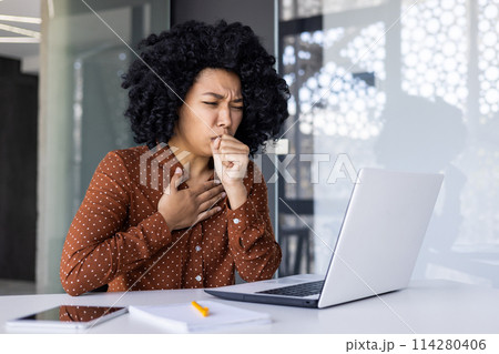 A young African American woman feeling unwell, coughing while working at her office desk. The image captures her discomfort and the healthcare concept in a workplace setting. 114280406