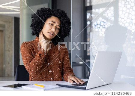 African-American woman experiencing discomfort and neck pain while working on a laptop in a modern office environment, signaling overuse or poor posture. 114280469