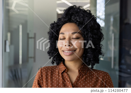 A portrait of a serene young woman enjoying a peaceful moment with closed eyes. Smiling gently, she stands indoors in a professional setting, radiating a calm and composed vibe. A portrait of a serene young woman enjoying a peaceful moment with closed eyes. Smiling gently, she stands indoors in a professional setting, radiating a calm and composed vibe. 114280477