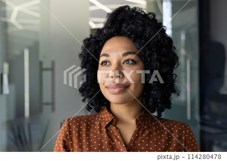 Portrait of a confident young African-American woman in a polka dot shirt, gazing away with a thoughtful look in a modern office setting. Portrait of a confident young African-American woman in a polka dot shirt, gazing away with a thoughtful look in a modern office setting. 114280478