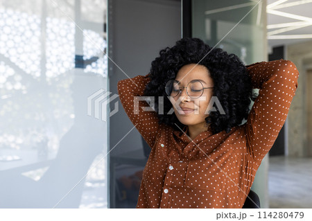 A young African American woman enjoys a peaceful moment in a bright, modern office. She is leaning back in her chair with her eyes closed, displaying a sense of relaxation and contentment. A young African American woman enjoys a peaceful moment in a bright, modern office. She is leaning back in her chair with her eyes closed, displaying a sense of relaxation and contentment. 114280479