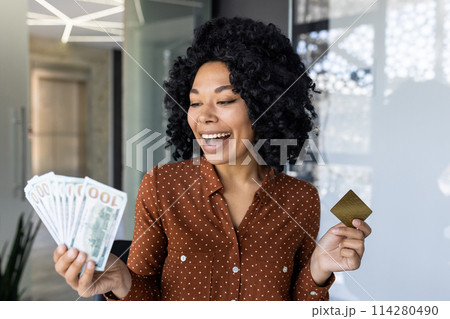 Joyful African American woman in a stylish polka dot blouse excitedly holds US dollar bills and a gold credit card in a modern office setting. 114280490