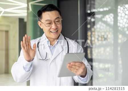 Friendly Asian mature male doctor engaging in a video call using a tablet. He's smiling and waving, positioned in a modern clinic setting. Friendly Asian mature male doctor engaging in a video call using a tablet. He's smiling and waving, positioned in a modern clinic setting. 114281515