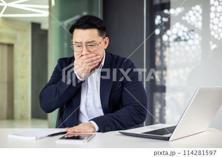 A middle-aged Asian businessman appears unwell, covering his mouth with his hand, while working at his laptop in a modern office setting. 114281597
