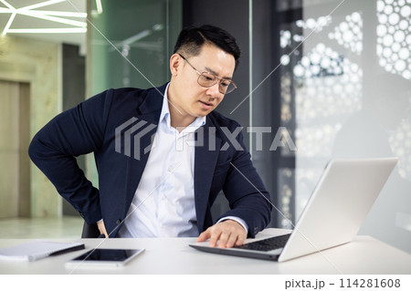Asian businessman in a modern office feeling a sharp pain in his back while working intensely on a laptop. Focus on health issues related to office work. 114281608