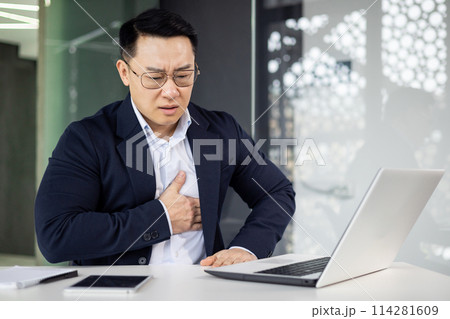An Asian businessman appears distressed, clutching his chest in pain while sitting at a desk in a modern office. He looks at his laptop screen with a worried expression. 114281609