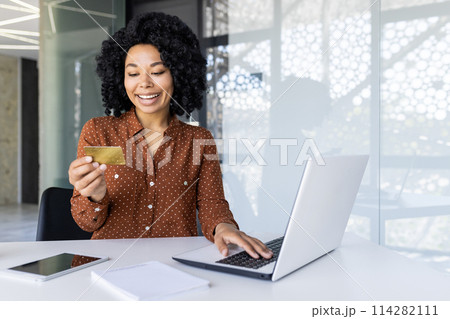 A cheerful young African American woman joyfully shopping online. She holds a credit card while using a laptop in a light, modern office space. 114282111