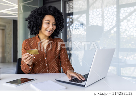 Cheerful young woman with curly hair using a credit card for an online purchase. Sitting at a desk with a laptop in a bright, modern office. 114282112