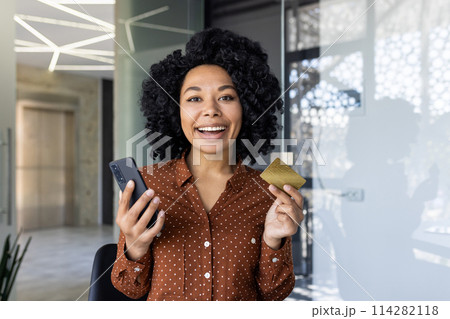 A cheerful African American businesswoman holds a credit card and smartphone in a modern office setting, symbolizing ecommerce and mobile banking. 114282118