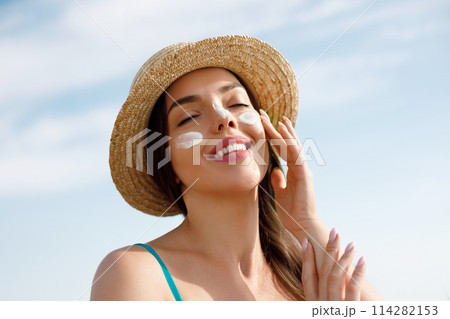 Beautiful young woman at beach applying sunscreen on face and looking at camera. Beauty girl applying suntan lotion at sea. Portrait of happy woman with healthy skin applying sunblock on cheek. 114282153
