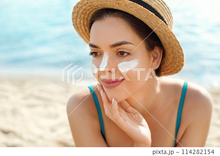 Beautiful woman applying cream sunscreen on a tanned face. Sunscreen. Skin and body care. The girl uses a sunscreen for her skin. Portrait of a female holding suntan lotion and moisturizing sunscreen. 114282154