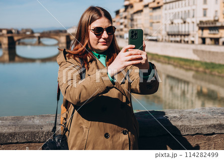 Attractive young female tourist is exploring new city. Redhead girl holding a smartphone on famous Old bridge in Florence. Traveling. Happy optimistic girl walking in city and makes selfie Attractive young female tourist is exploring new city. Redhead girl holding a smartphone on famous Old bridge in Florence. Traveling. Happy optimistic girl walking in city and makes selfie 114282499