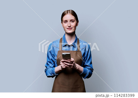 Young female worker in an apron using smartphone on gray studio background 114283399