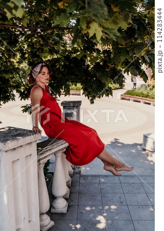 A pretty woman in a red silk dress and a bandage on her head smiles against the background of the leaves of a tree. She is leaning on the coop and looking into the camera. Vertical photo. 114283444