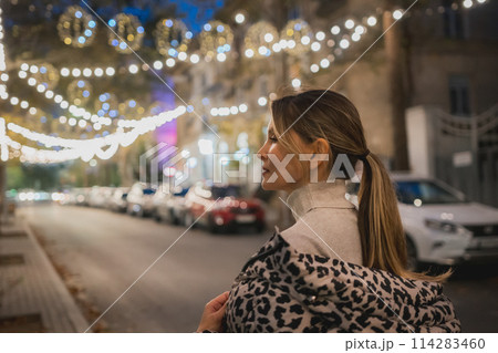 A woman wearing a leopard print coat stands on a street at night. The street is lit up with Christmas lights, creating a festive atmosphere. There are several cars parked along the street. A woman wearing a leopard print coat stands on a street at night. The street is lit up with Christmas lights, creating a festive atmosphere. There are several cars parked along the street. 114283460
