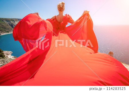 woman sea red dress. Blonde with long hair on a sunny seashore in a red flowing dress, back view, silk fabric waving in the wind. Against the backdrop of the blue sky and mountains on the seashore. 114283525
