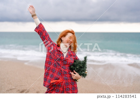 Sea Lady in plaid shirt with a christmas tree in her hands enjoys beach. Coastal area. Christmas, New Year holidays concep 114283541