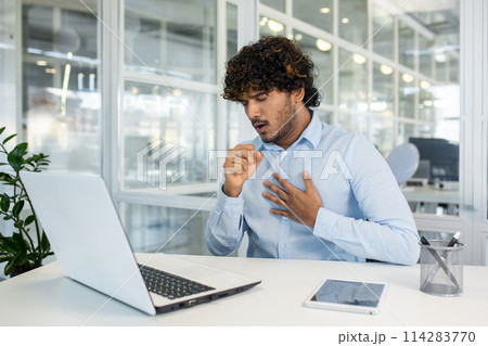A young man in a light blue shirt feels a sudden cough while working on his laptop in a modern office setting. Concern and discomfort are evident as he addresses his health at work. A young man in a light blue shirt feels a sudden cough while working on his laptop in a modern office setting. Concern and discomfort are evident as he addresses his health at work. 114283770