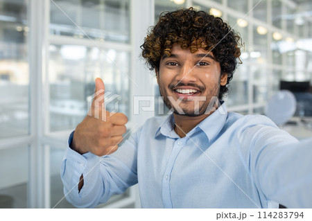 Confident young man with curly hair showing a thumbs up while taking a selfie at a bright, well-lit office setting. Positive and professional vibe. 114283794