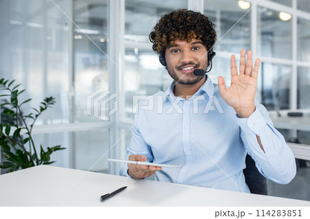 A cheerful call center representative waves at the camera, wearing a headset and holding a digital tablet in a modern office setting. 114283851