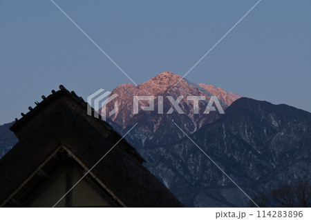 茅葺き屋根の向うに朝焼けの南アルプス甲斐駒ケ岳 114283896