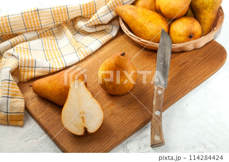 Cutting board with whole and halves pears, wicker basket and yellow kitchen towel on white wooden background.. 114284424